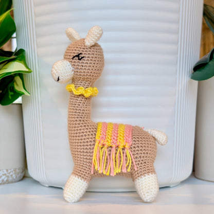 Side view of Crocheted vicuna llama beige plush with a yellow and pink saddle and a yellow necklace