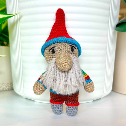 A handmade crocheted gnome doll with a red hat, blue trim, beige face and beard, white beard, and rainbow-striped sleeves, standing in front of a white ribbed container.