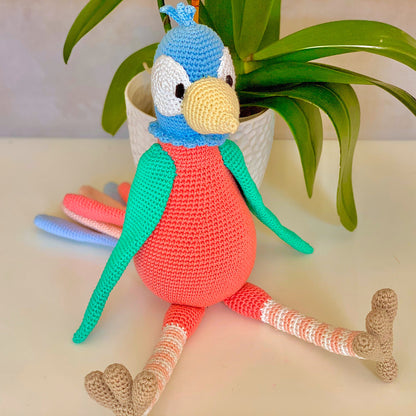 Big crocheted bird with coral body, blue head, turquoise green wings, and multi-colored tail feathers sitting on a table. 