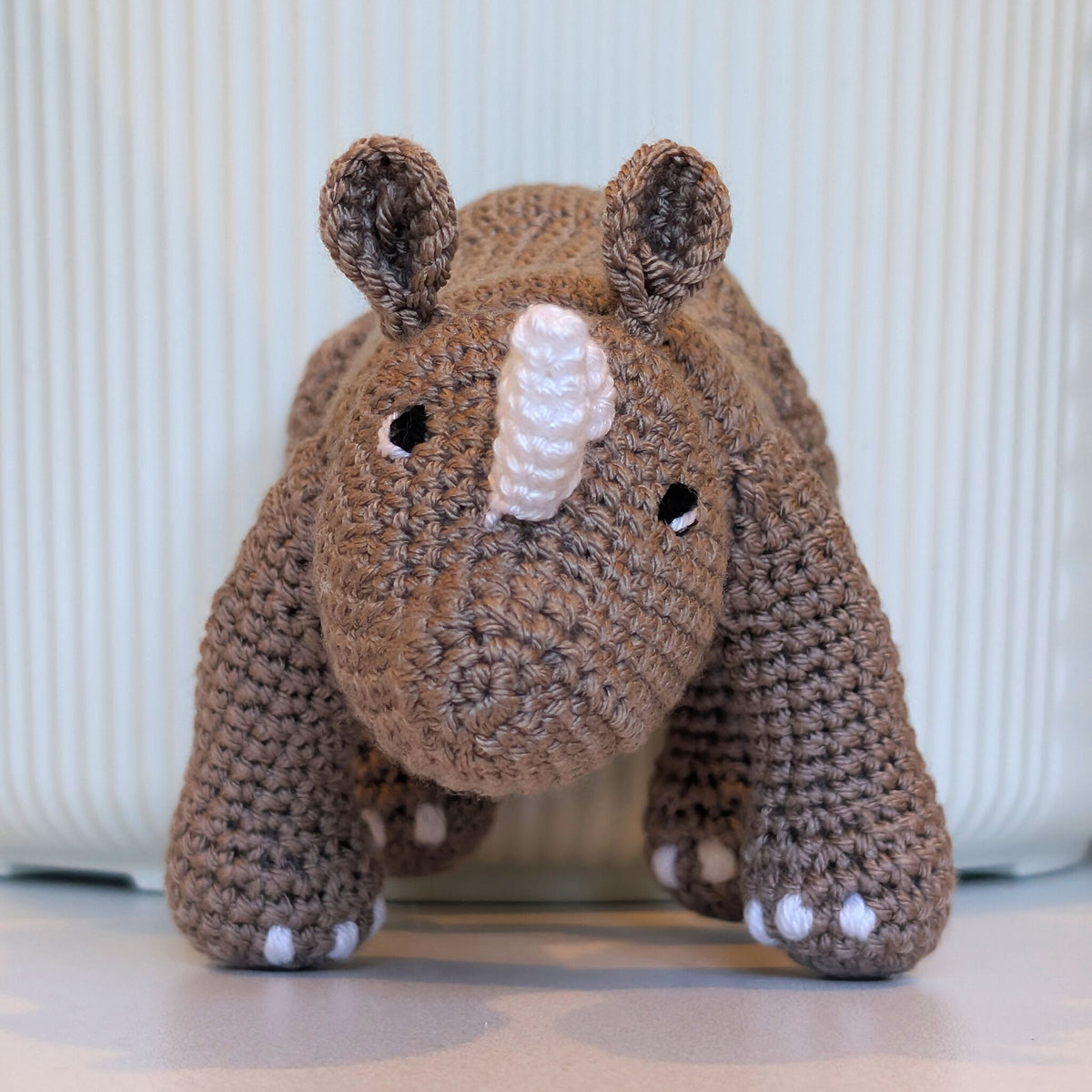 A handmade crocheted rhinoceros toy in taupe-brown yarn with white accents on its horns and feet, displayed on a light surface in front of a ribbed white ceramic container.