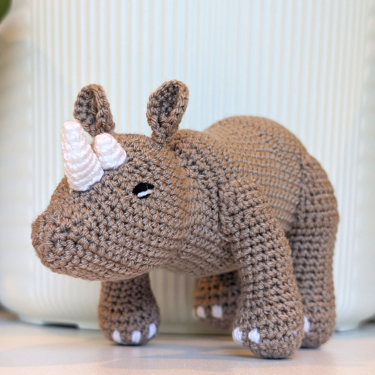 A crocheted rhinoceros toy in warm brown yarn with white accents on its horn, feet, and ears, standing against a soft-focus light green background.