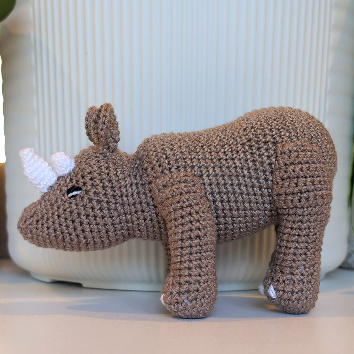 A handmade crocheted rhinoceros toy in taupe-brown yarn with white accents on its horns and feet, displayed on a light surface in front of a ribbed white ceramic container.