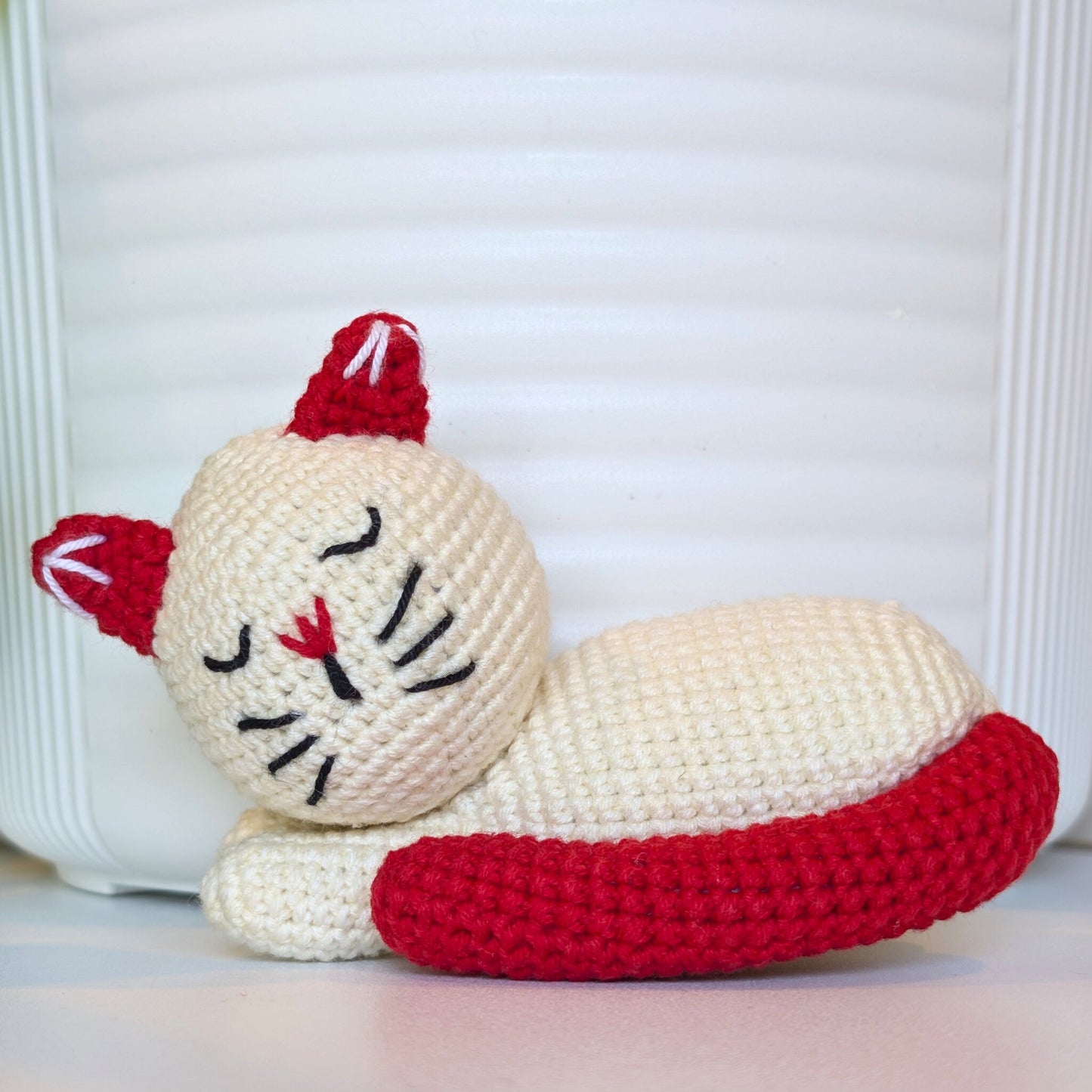 A crocheted sleeping cat toy in cream and red, positioned against a white ribbed background.