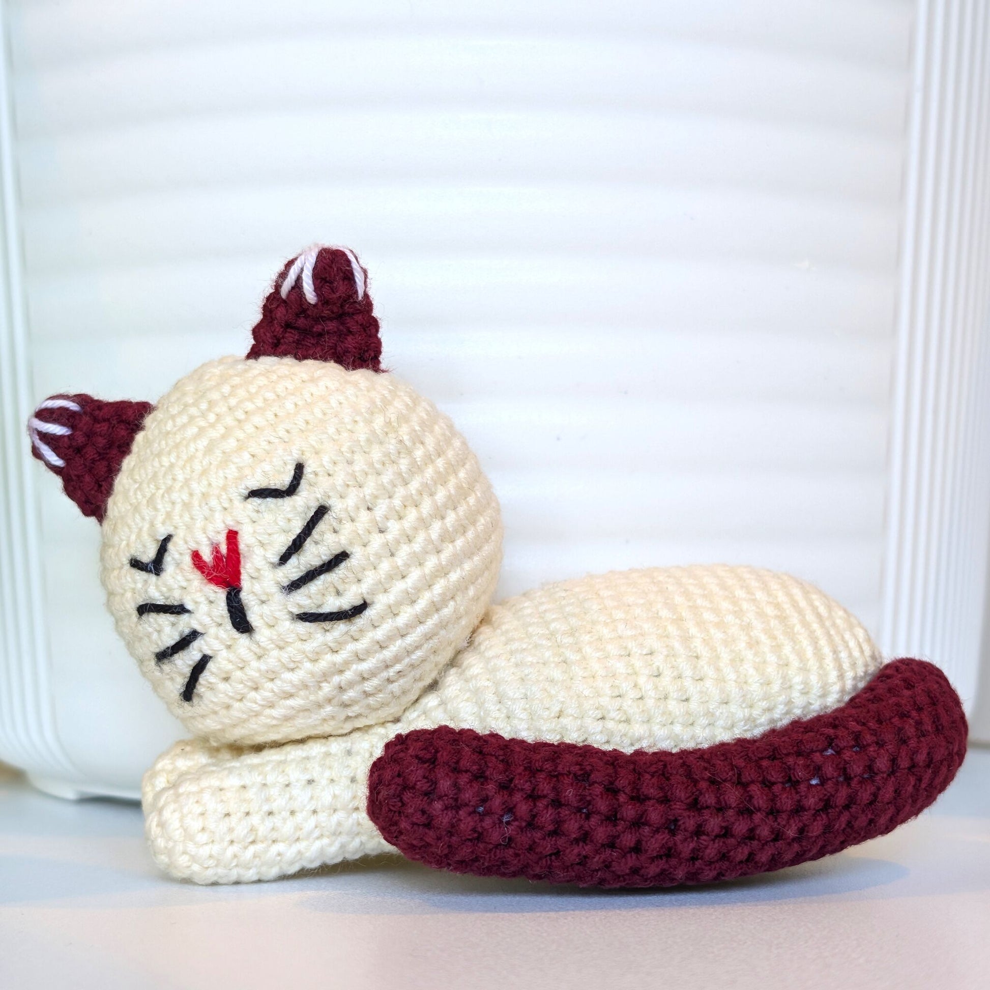 A crocheted sleeping cat toy in cream white with dark burgundy ears, tail, and whiskers, plus a small red stitched nose, posed against a white slatted background.