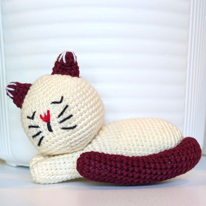 A crocheted sleeping cat toy in cream white with dark burgundy ears, tail, and whiskers, plus a small red stitched nose, posed against a white slatted background.
