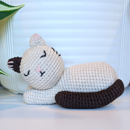 A crocheted sleeping cat toy in off-white with dark brown tail, and ears, resting on a white surface in front of a white ribbed background.