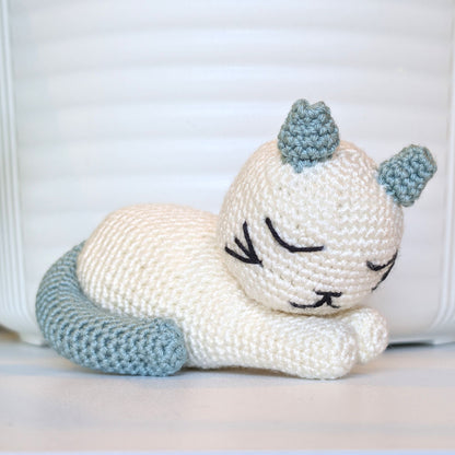 A crocheted sleeping cat toy in off-white with light blue ears, tail, and back, resting on a white surface against a white background.