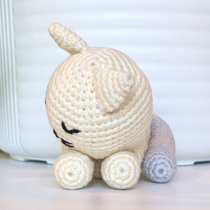 A handmade crocheted amigurumi toy resembling a small sleeping cat or animal, made from cream-colored yarn with a light gray tail, sitting on a white surface against a white background.