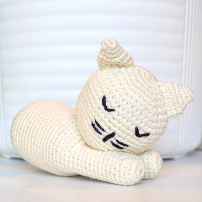 A cream-colored crocheted cat toy lying down, featuring black embroidered eyes, nose, and whiskers, set against a white ribbed background.