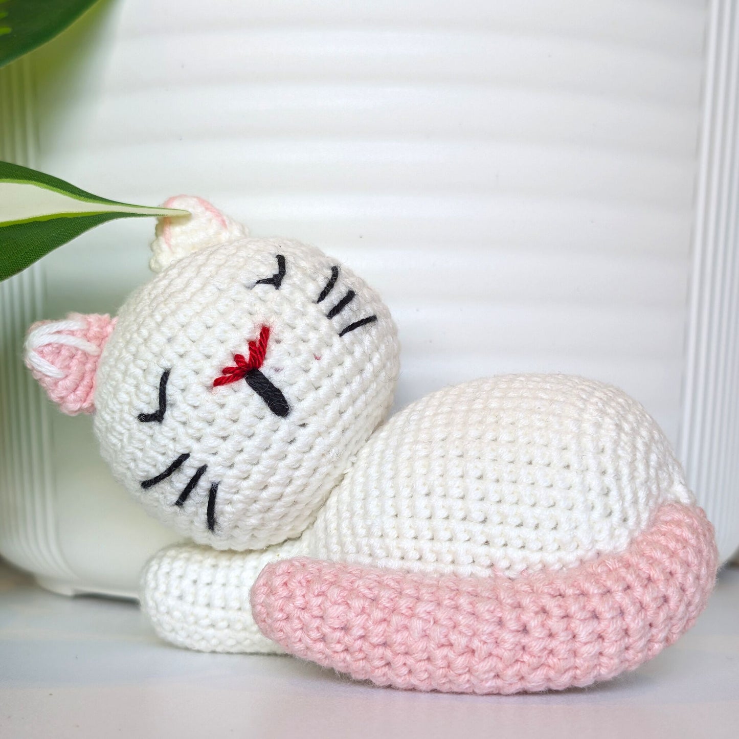 A handmade crocheted cat toy in white and light pink, shown lying down with embroidered black facial features and a small red nose detail, set against a white ribbed background with a green leaf in the corner.