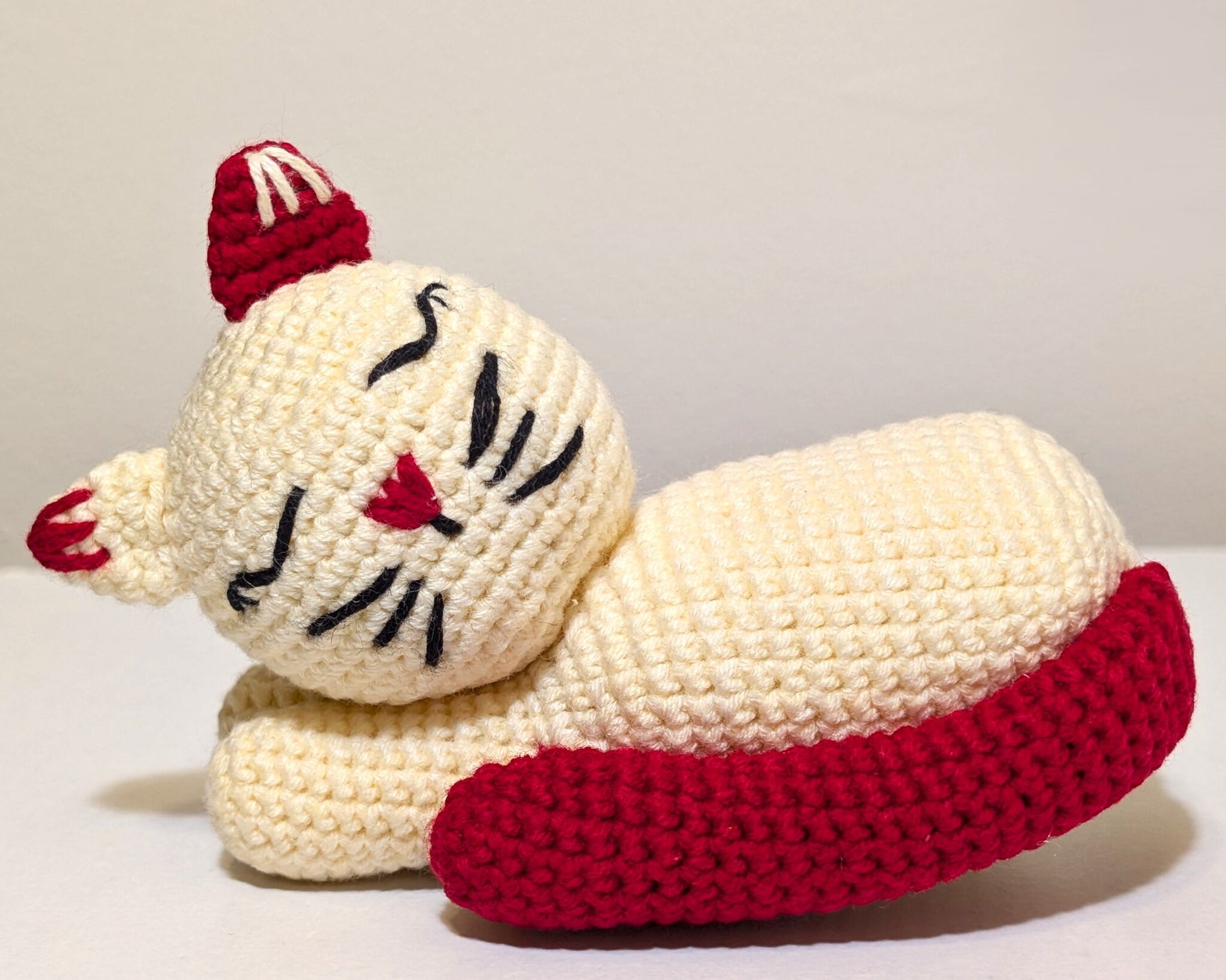A handmade crocheted cat toy in cream and red, lying down on a plain light background.