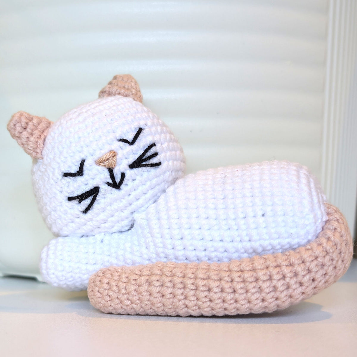 A crocheted sleeping cat toy in white with light beige ears, tail, and nose, and black embroidered eyes and whiskers, resting on a light surface.
