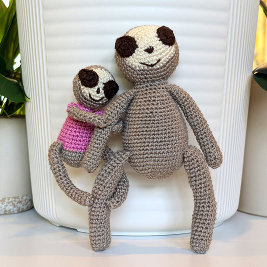 Mom and baby crocheted sloths on a light surface, baby wearing very light orchid top