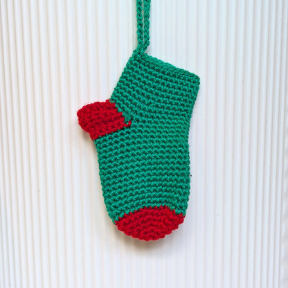 A green and red hand-crocheted Christmas stocking ornament hanging against a white ribbed background.