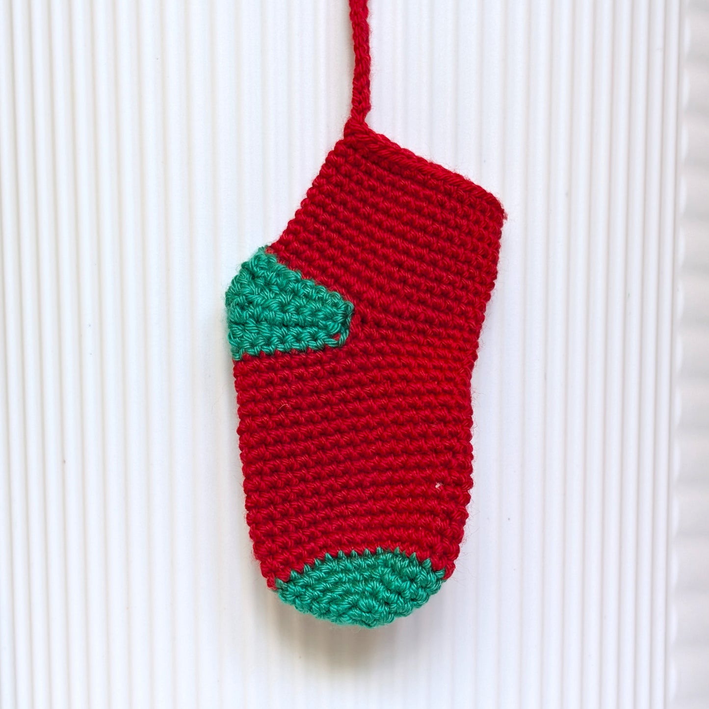 A hand-crocheted Christmas stocking ornament in red and green, hanging against a white ribbed background.