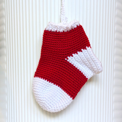 A small crocheted Christmas stocking ornament in red and white, hanging against a white ribbed background.