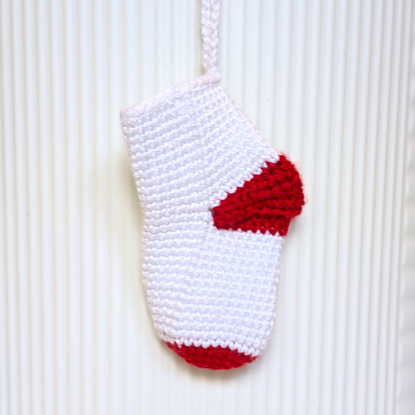 A white crocheted sock ornament with a red heel and toe, hanging against a white ribbed background.