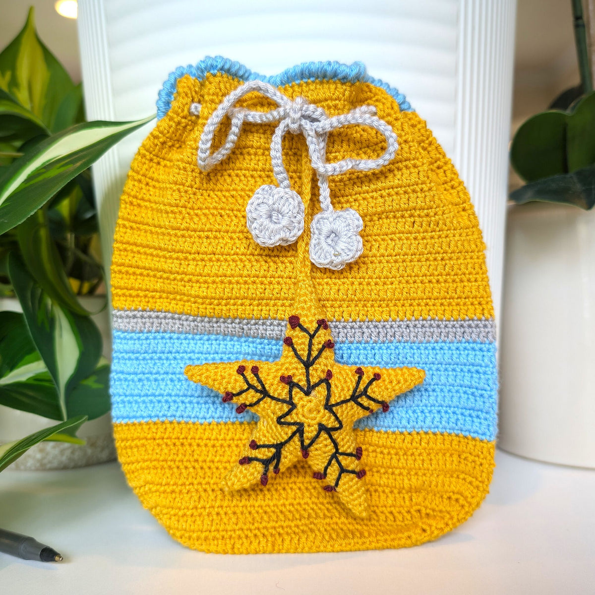 A handmade crocheted drawstring pouch in mustard yellow and light blue, featuring a gray and white drawstring with flower charms and a gray horizontal stripe. A yellow starburst with a dark red snowflake design is embroidered on the front.