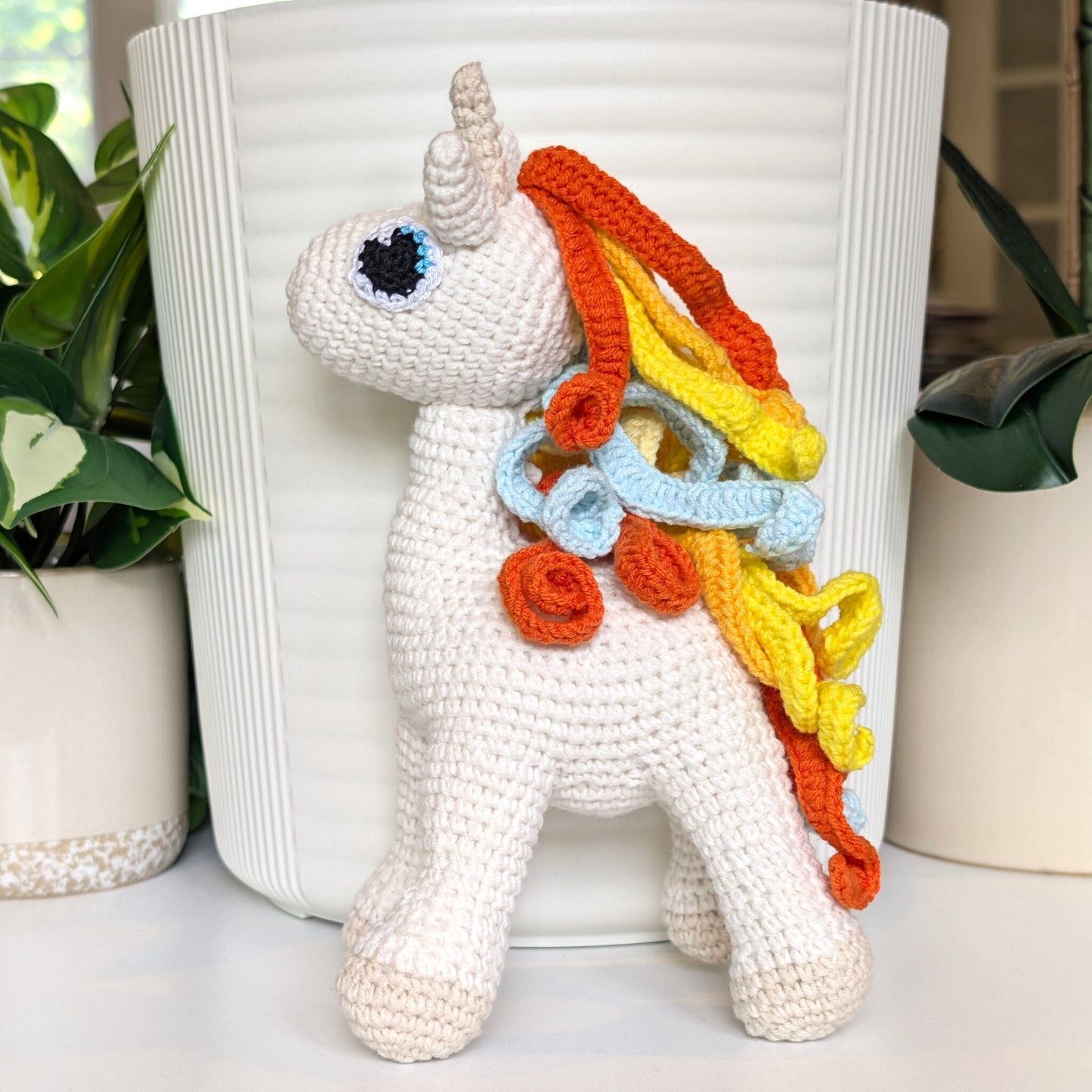 Crocheted plush unicorn toy with colorful rainbow orange and yellow hair in front of a white pot.