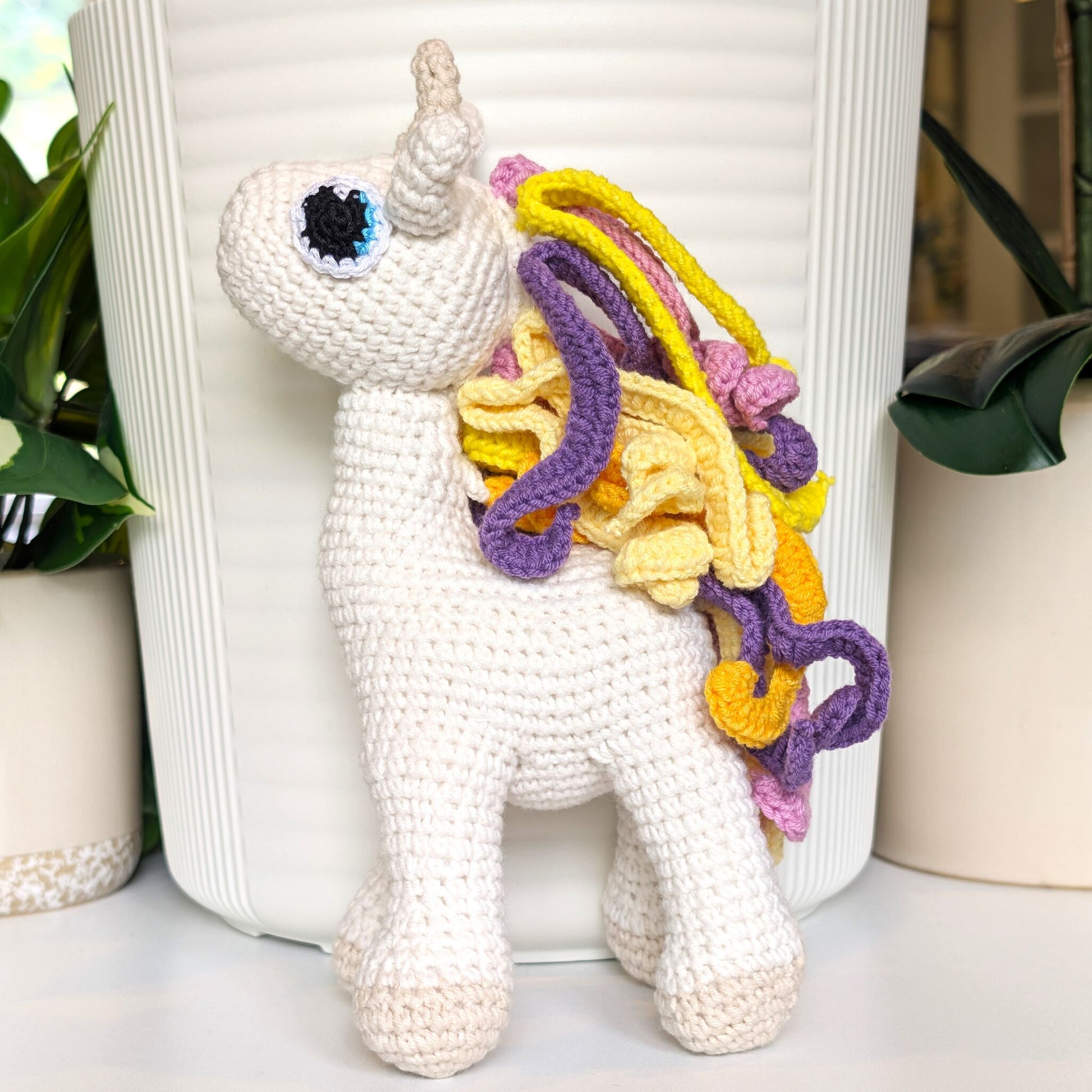 White crochet plush unicorn with colorful mane in purple, yellow and pink colors against a white background