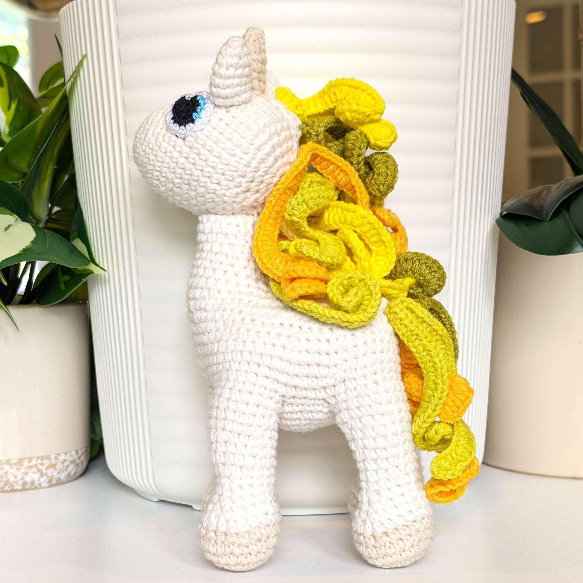 White crocheted plush unicorn toy with a yellow mane and tail in front of a white vase.