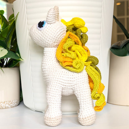 White crocheted plush unicorn toy with a yellow mane and tail in front of a white vase.