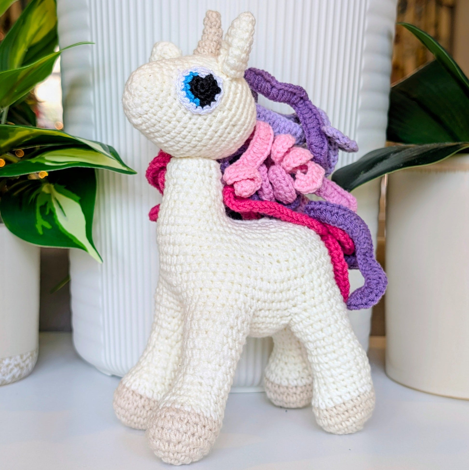 Crocheted unicorn toy with colorful pink and purple mane and tail on a white surface with plants in the background