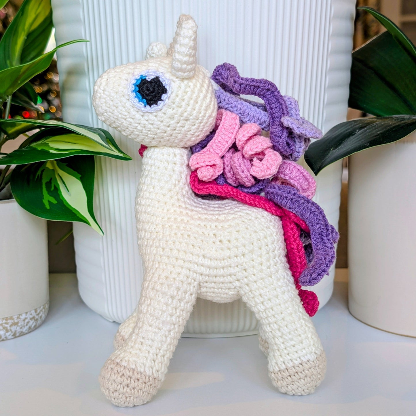 Crocheted soft unicorn toy with a colorful pink and purple mane in front of plants