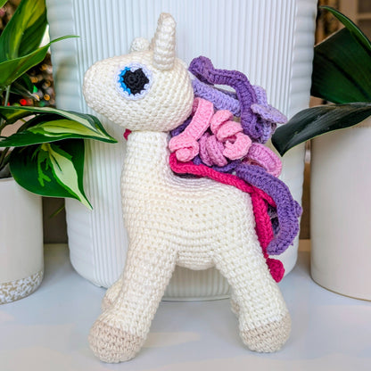 Crocheted soft unicorn toy with a colorful pink and purple mane in front of plants