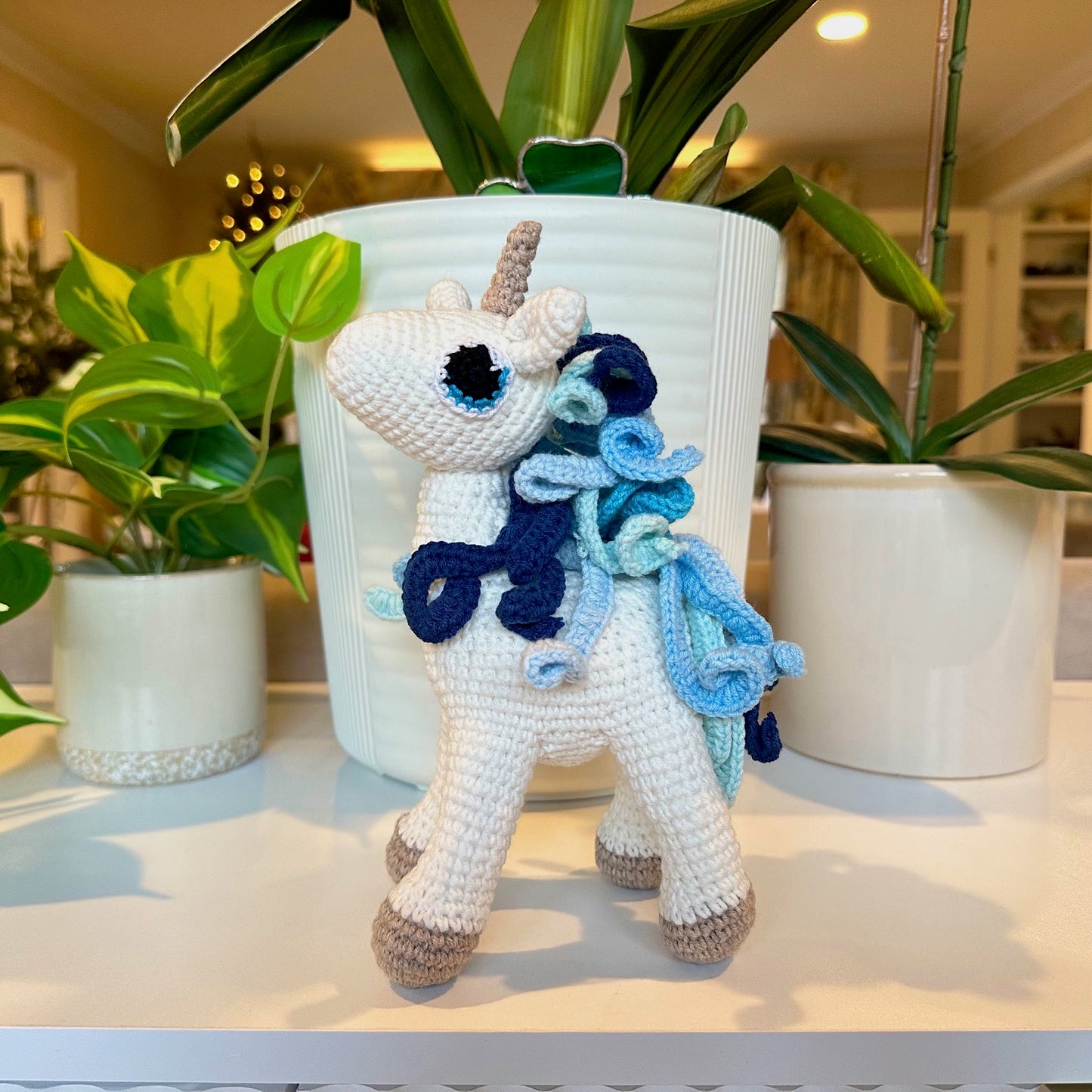 Crocheted unicorn toy with blue mane and tail on a surface with plants in the background