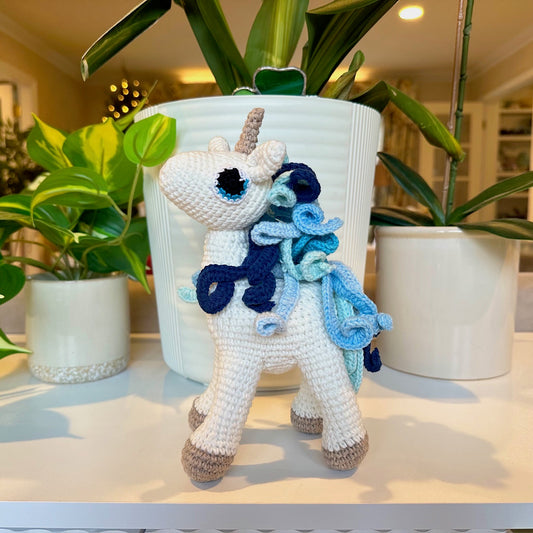 Crocheted unicorn toy with blue mane and tail on a surface with plants in the background