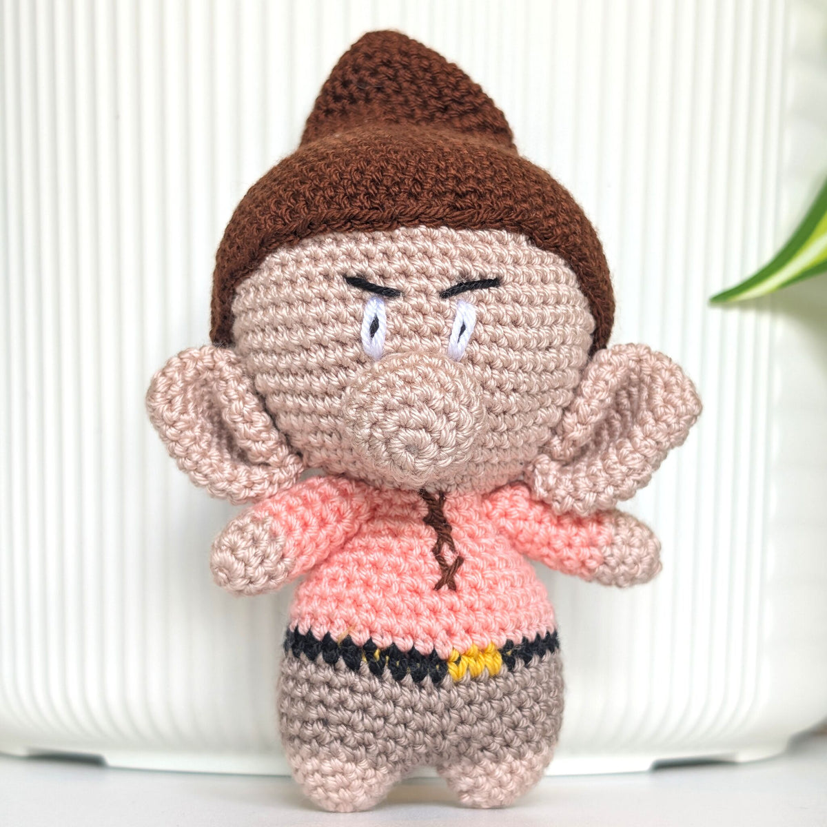 A crocheted amigurumi doll with a brown pointed hat, beige skin, large ears, a pink top, and gray pants, standing against a white ribbed background.