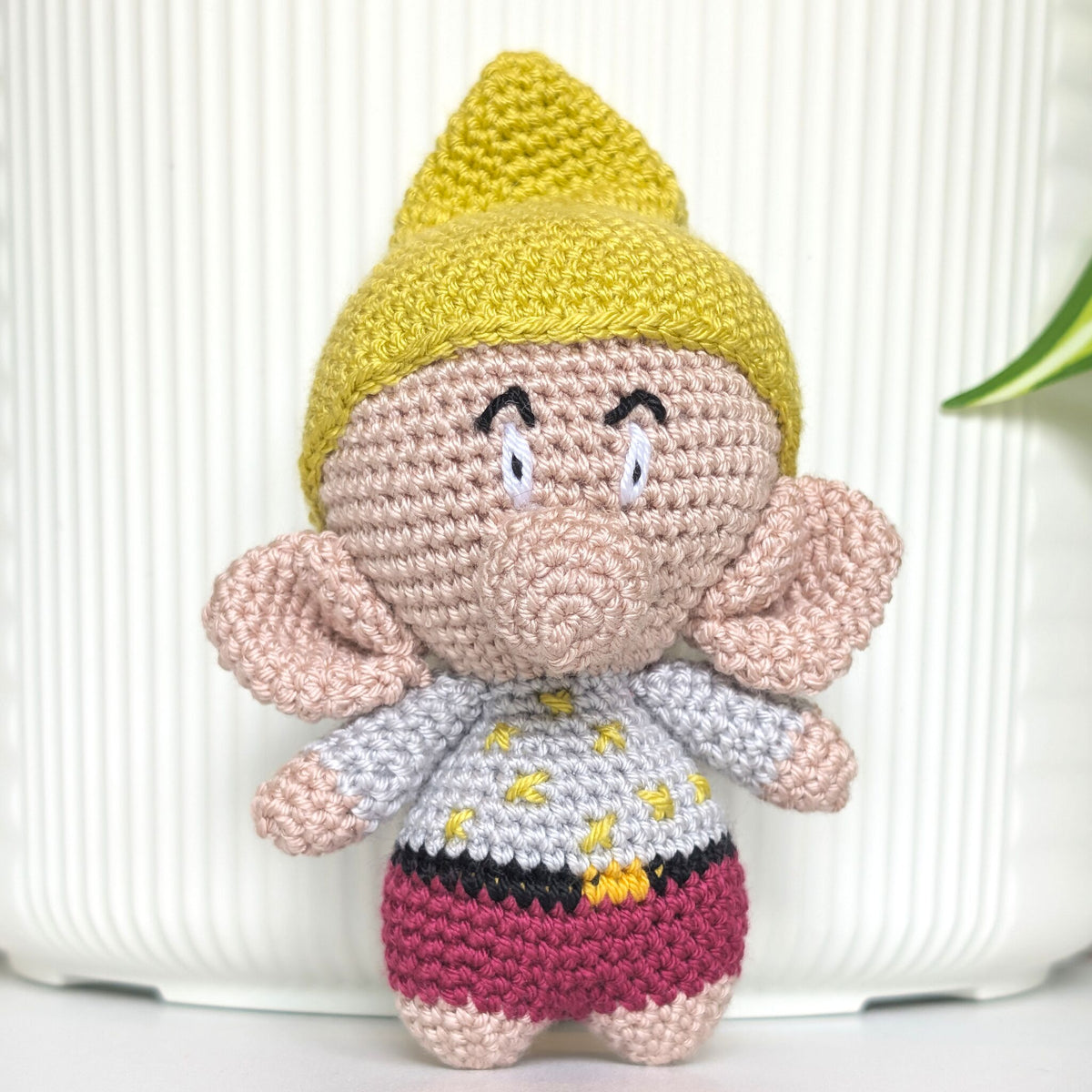 A handmade crocheted amigurumi doll of a gnome character, featuring a chartreuse hat, beige face and ears, a white shirt with chartreuse dot, and a magenta-red shorts, set against a white ribbed background.