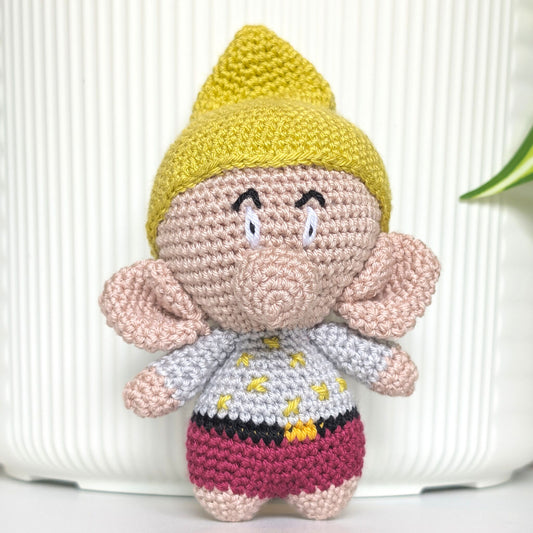 A handmade crocheted amigurumi doll of a gnome character, featuring a chartreuse hat, beige face and ears, a white shirt with chartreuse dot, and a magenta-red shorts, set against a white ribbed background.