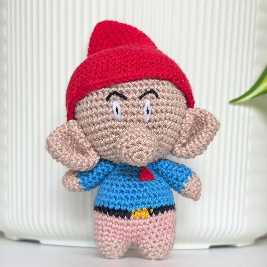 A handmade crocheted amigurumi doll resembling a gnome character, featuring a rose hat, beige skin, a blue shirt, and pink pants, set against a white ribbed background.