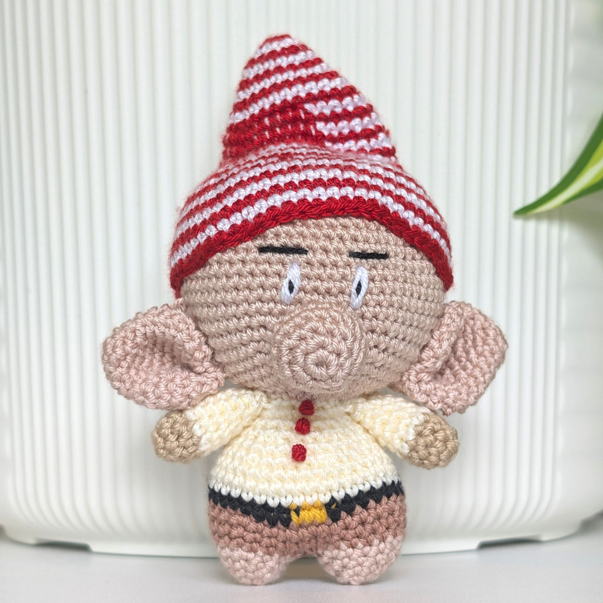A handmade crocheted gnome doll with a red and white striped hat, beige skin, and a cream-colored shirt with red buttons, standing in front of a white ribbed background.