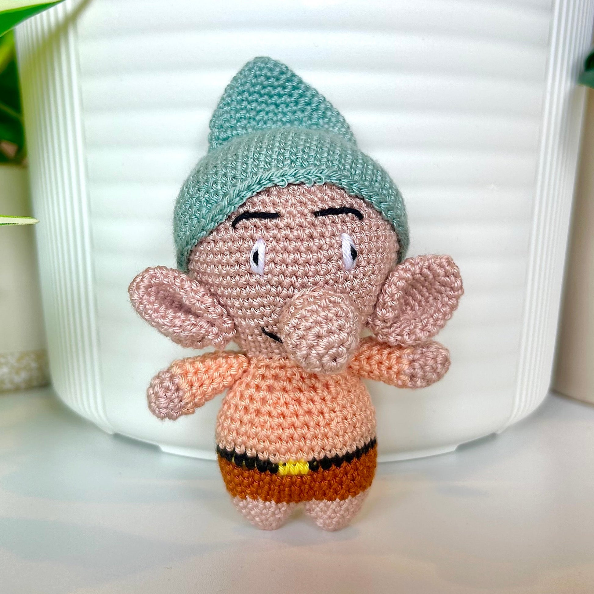 Crocheted gnome toy with a seafoam blue green hat in front of a white pot.