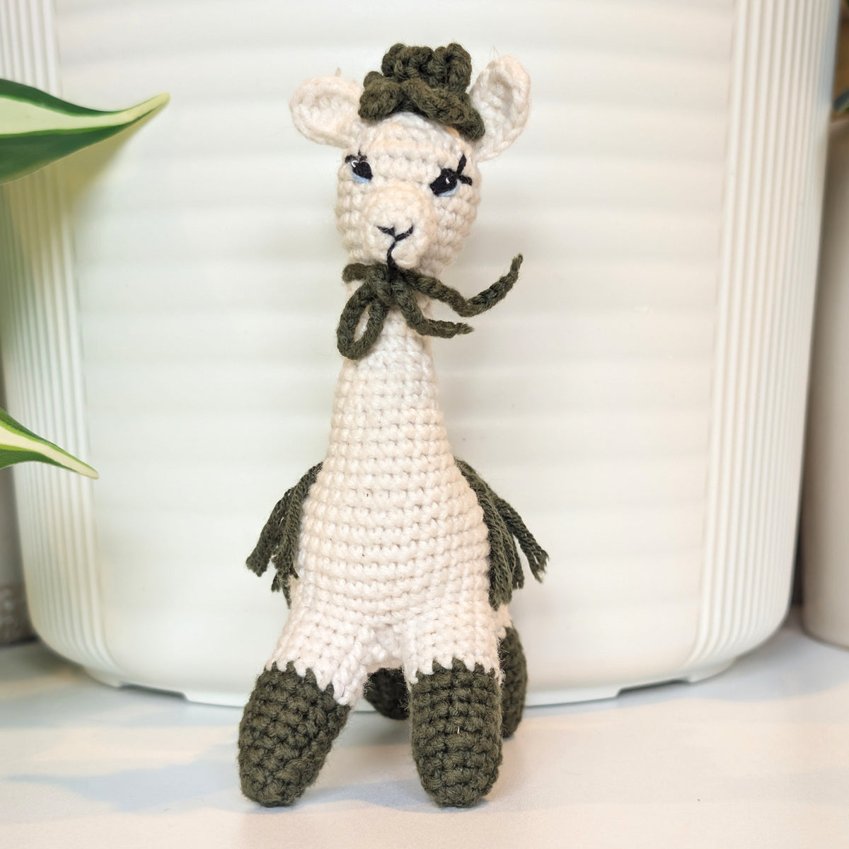 A crocheted llama or alpaca doll in off-white with dark olive green accents, including a hat, bow, tail, and hooves, standing in front of a white ribbed ceramic pot.