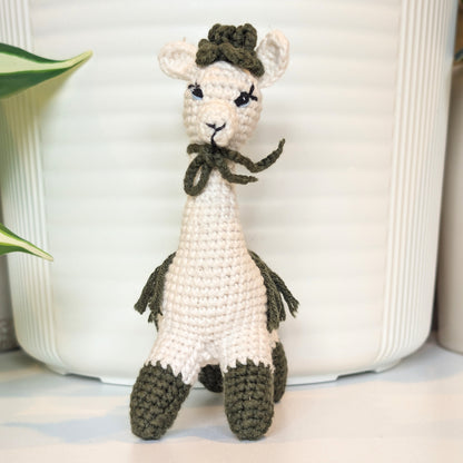 A crocheted llama or alpaca doll in off-white with dark olive green accents, including a hat, bow, tail, and hooves, standing in front of a white ribbed ceramic pot.