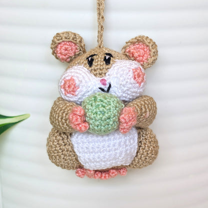 A crocheted hanging hamster ornament in tan, white, pink, and green yarn, holding a small green ball, photographed against a white ribbed background.