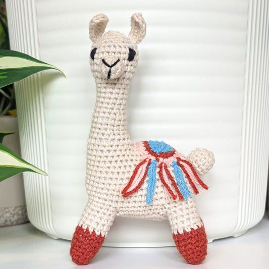 A crocheted llama plush toy with a cream-colored body, red hooves, and a blue and red saddle detail, standing in front of a white ribbed planter pot with a green plant leaf on the left.
