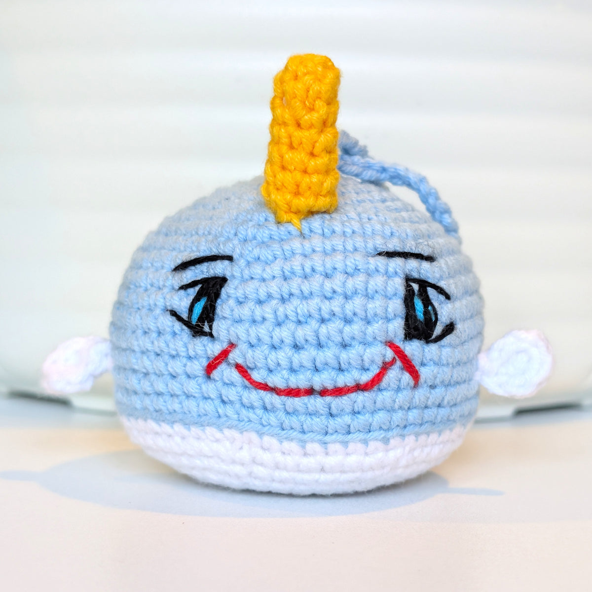 A handmade crocheted plush toy narwhal with a yellow horn, embroidered eyes, a red smile, photographed against a soft white background.