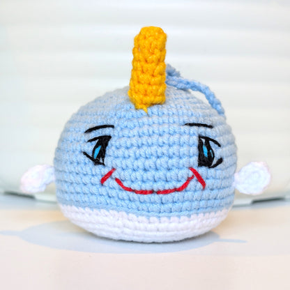 A handmade crocheted plush toy narwhal with a yellow horn, embroidered eyes, a red smile, photographed against a soft white background.
