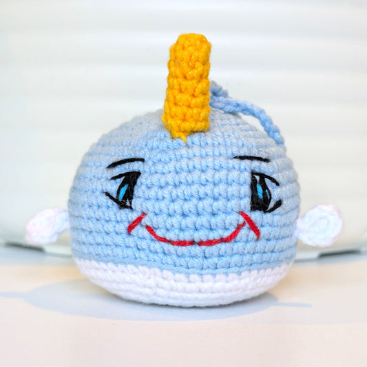 A handmade crocheted plush toy narwhal with a yellow horn, embroidered eyes, a red smile, photographed against a soft white background.