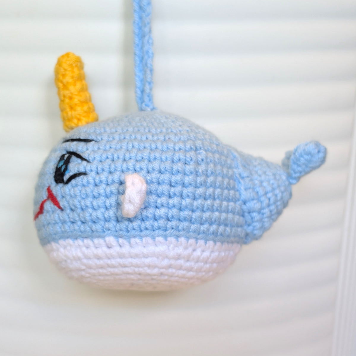A crocheted narwhal ornament or bag charm in light blue and white yarn with a yellow horn, hanging against a white slatted background.