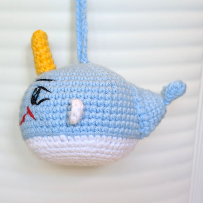 A crocheted narwhal ornament or bag charm in light blue and white yarn with a yellow horn, hanging against a white slatted background.