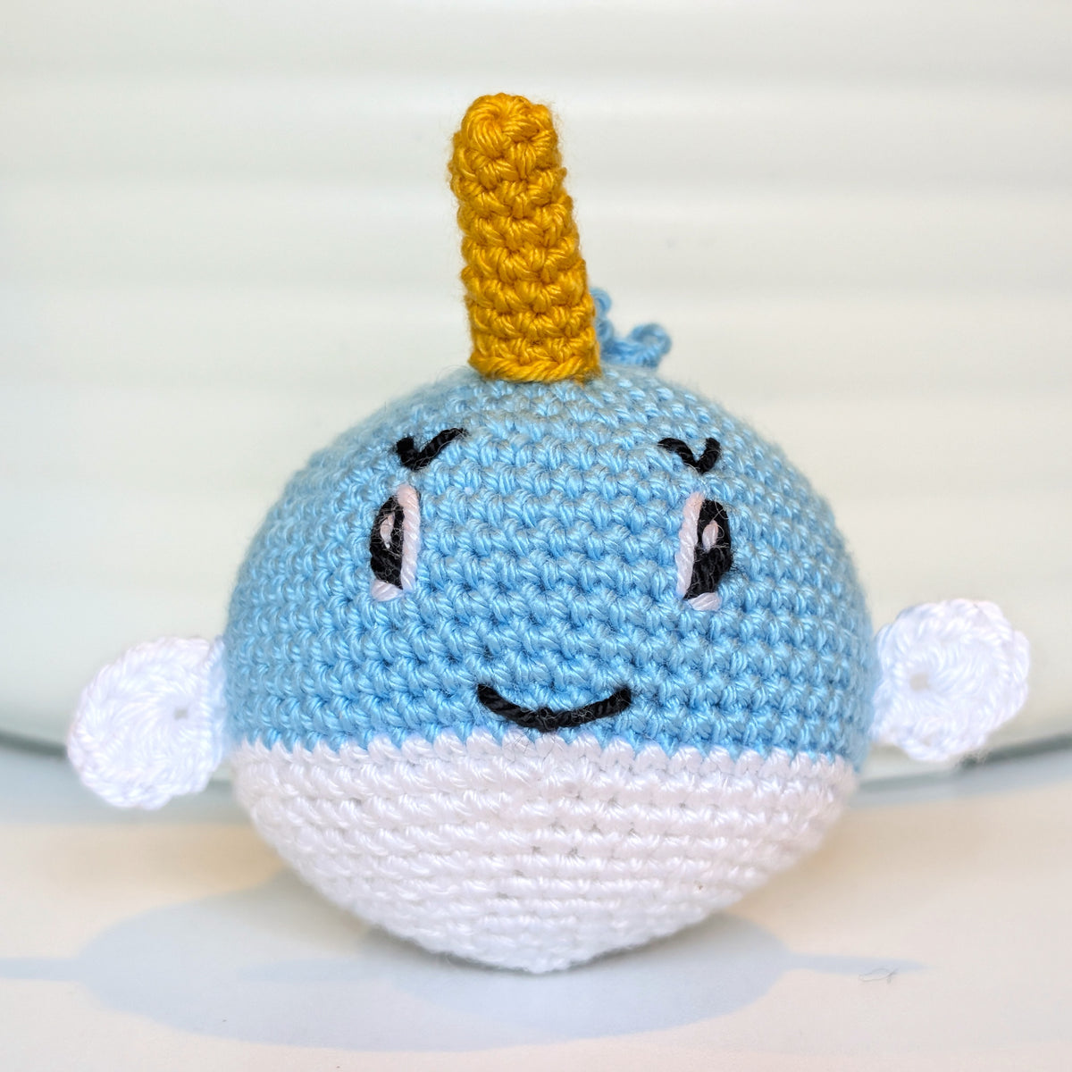 A handmade crocheted narwhal toy with a caribbean blue and white body, a yellow horn, and embroidered facial features, set against a soft, light-colored background.