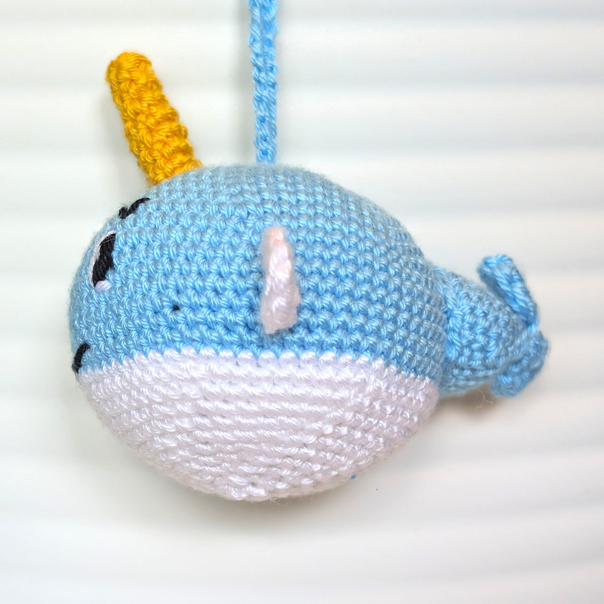 A handmade crocheted narwhal toy with a caribbean blue body, white belly, yellow horn, and tusks, hanging against a white paneled background.