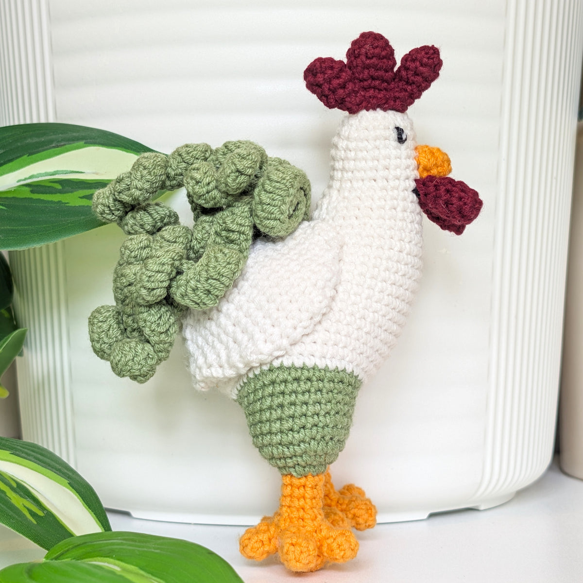 A crocheted rooster figurine with a white body, green tail feathers, orange beak and feet, and a dark red comb, posed in front of a white ribbed container with green leaves in the foreground.