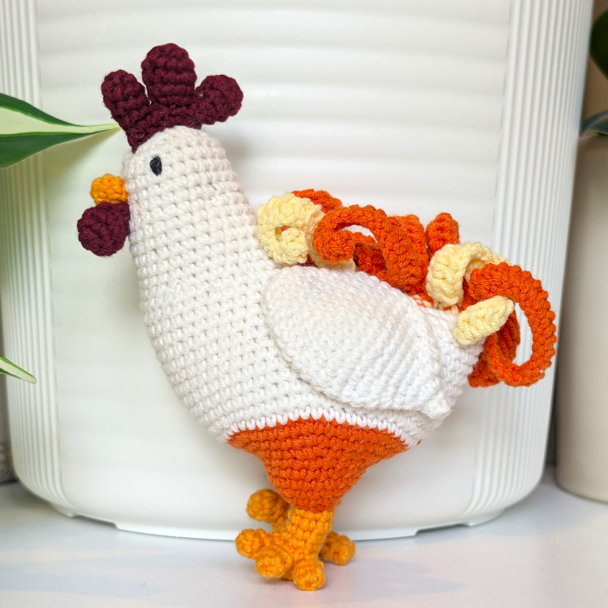 A crocheted toy rooster with a white body, orange legs and tail, and a dark burgundy comb and wattle, posed in front of a white ribbed ceramic pot.